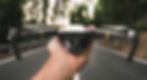 Close-up of a hand holding a drone with its rotors and camera visible, set against the backdrop of a road stretching into the distance. The focus is on the drone, emphasizing its compact size and intricate design, while the road behind it fades slightly, creating a sense of depth and context.