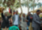 Five men hold books and smile outdoors under trees. One raises a fist, another gives a thumbs-up. Joyful mood, people in the background.
