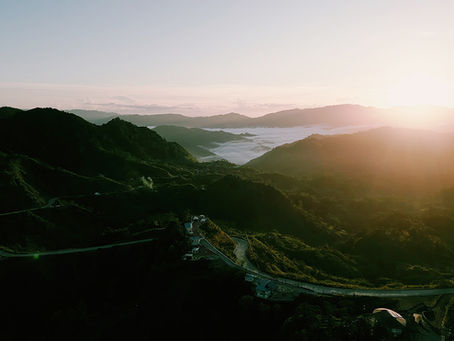 Sunrise over lush green mountains and winding roads, with misty valleys and a serene sky, creating a peaceful and tranquil scene.