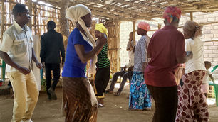 People dance energetically in a rustic open structure with wooden walls. They wear colorful headscarves, skirts, and shirts, with a joyful mood.