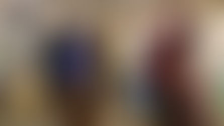 People dance energetically in a rustic open structure with wooden walls. They wear colorful headscarves, skirts, and shirts, with a joyful mood.