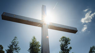 Wooden cross against a clear blue sky with sun rays, trees below, and a few clouds floating, creating a serene and peaceful mood.
