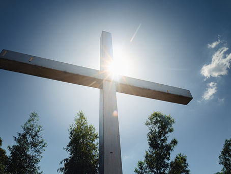 Wooden cross against a clear blue sky with sun rays, trees below, and a few clouds floating, creating a serene and peaceful mood.