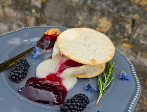 Oozing cheese and red preserve sandwiched between 2 crackers on grey plates, with gold spoon, blackberries, rosemary