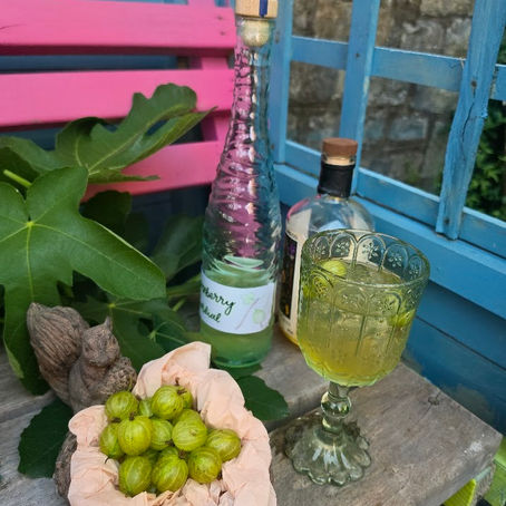 Green drink in an ornate glass beside bottles and a bowl of gooseberries on a blue-pink garden bench.
