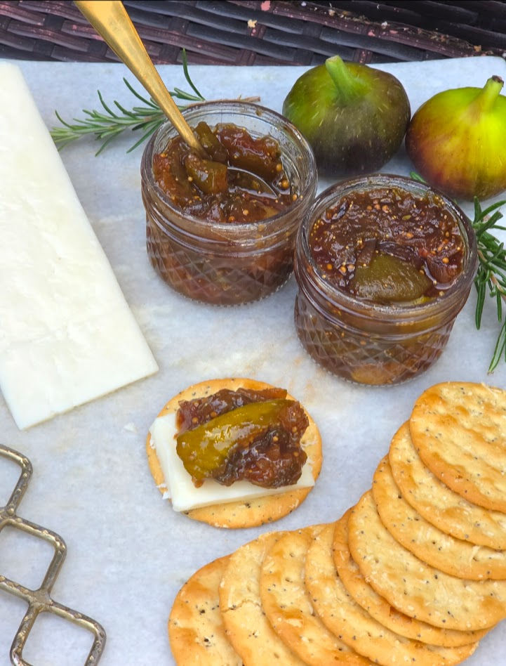 Fig chutney in jars with spoon, spread on crackers with cheese; fresh figs and rosemary on parchment, appetizer close-up.