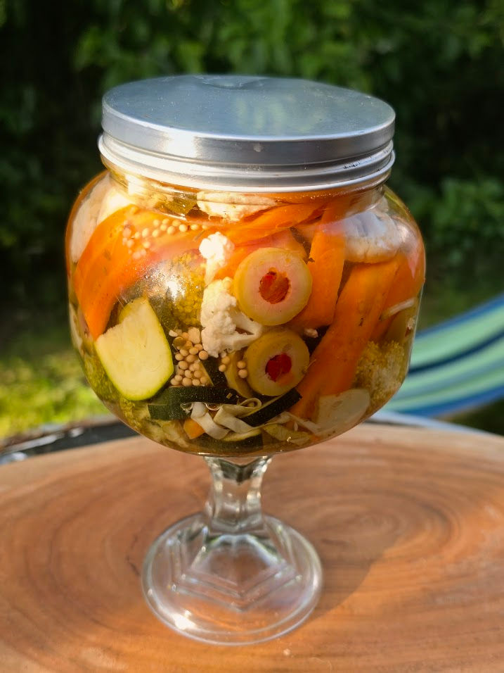 An image of a glass jar with a stem and a silver lid. Its filled with fresh, colourful vibrant sliced vegetables in a brine. Set on a wooden board in a leafy garden