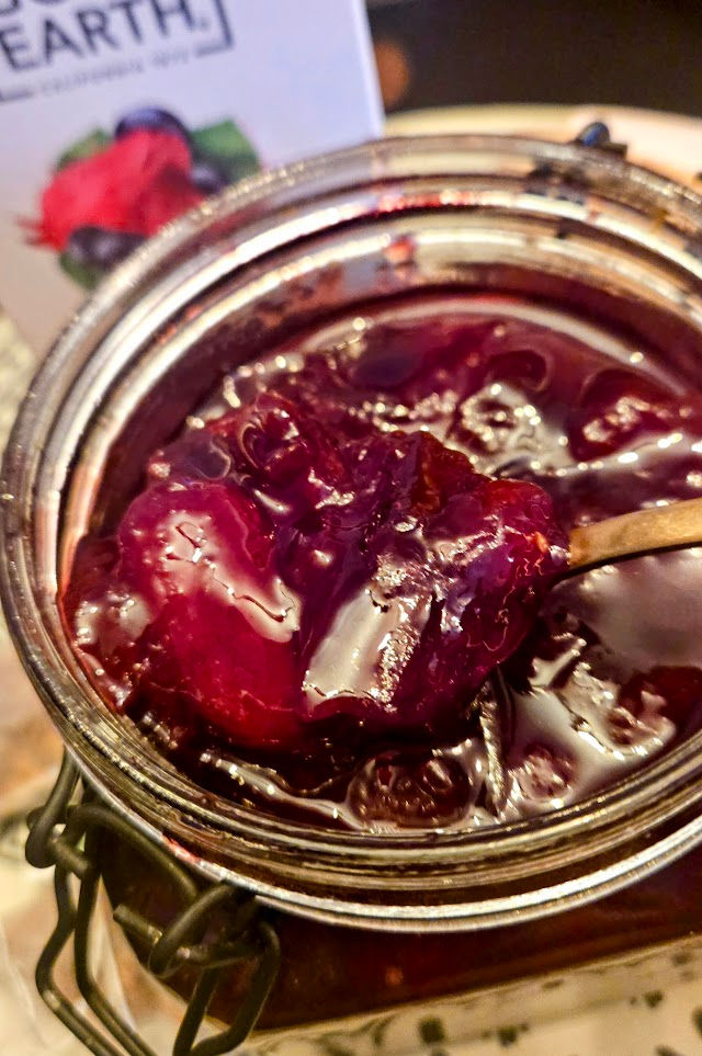 Open jar of dark red jam with a spoon inside. Background shows part of a box with the word "EARTH" visible. Warm, cozy setting.
