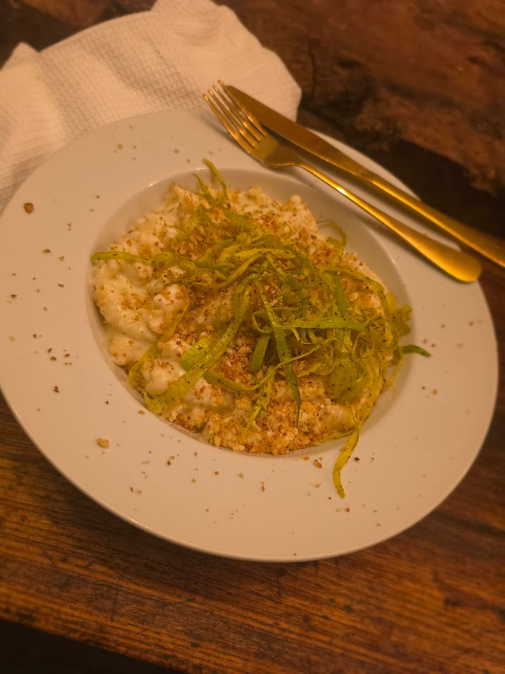 A plate of creamy risotto topped with shredded green garnish and crumbled breadcrumbs on a rustic wooden table. Gold utensils and a white napkin beside.