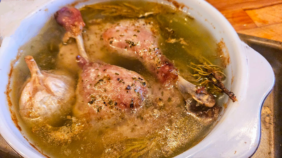 Cooked duck legs and garlic bulbs in a white dish with broth, garnished with herbs. Set on a wooden surface, creating a rustic feel.