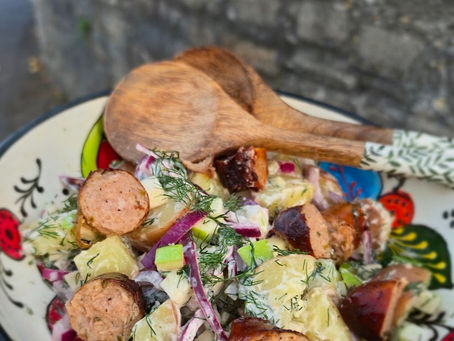 Colorful potato salad with sausage slices, red onion and dill in a patterned bowl, with wooden spoons on a stone ledge.