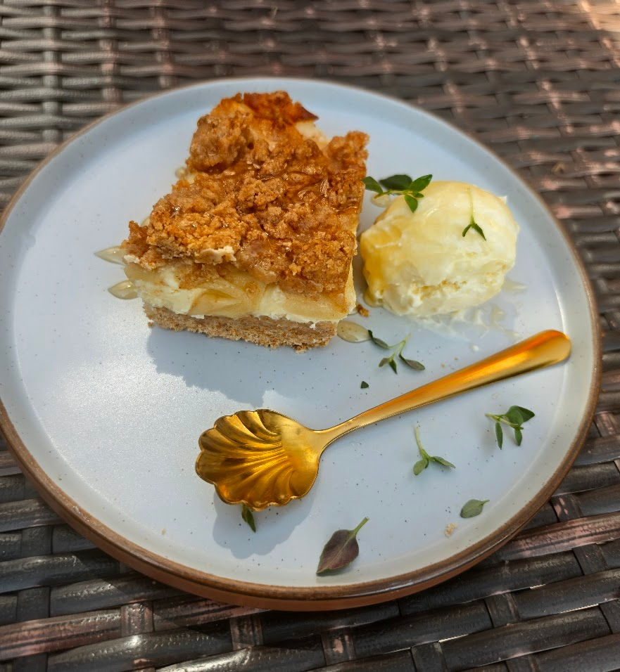 Slice of crumb-topped pie with a scoop of vanilla ice cream and gold spoon on a white plate atop a wicker table.