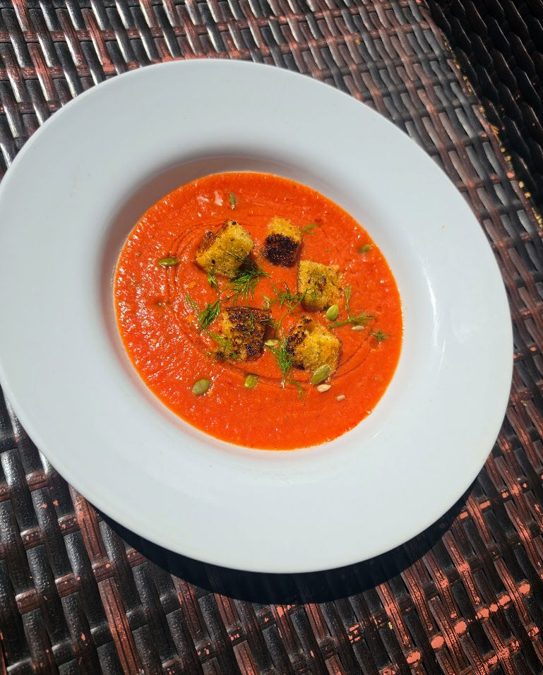 White bowl of red tomato soup topped with croutons and herbs on a woven dark table.