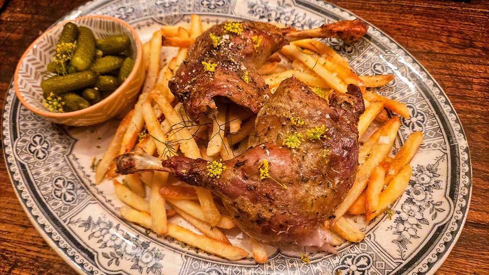 Roasted duck legs with herbs on fries, garnished with yellow flowers. Pickles in a bowl. Blue-patterned plate on a wooden table.