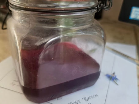 Glass jar with purple liquid and wooden lid sits on a note reading TEST BATCH with a heart on a kitchen counter.