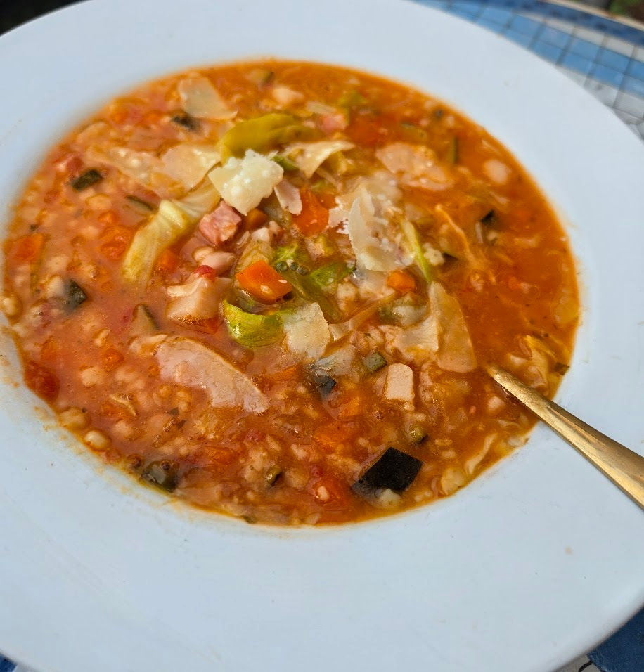 A hearty bowl of minestrone soup in a white bowl. Lots of fresh vibrant veggies and bacon pieces in a tomato broth, finished with parmesan shavings