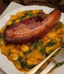 Plate of roasted pork belly over creamy beans and greens, with fork and knife on a white plate, warm hearty meal