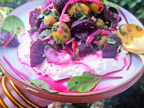 Vibrant beet and olive salad on a plate with creamy base, garnished with greens and red onions. Outdoor setting with leafy background.