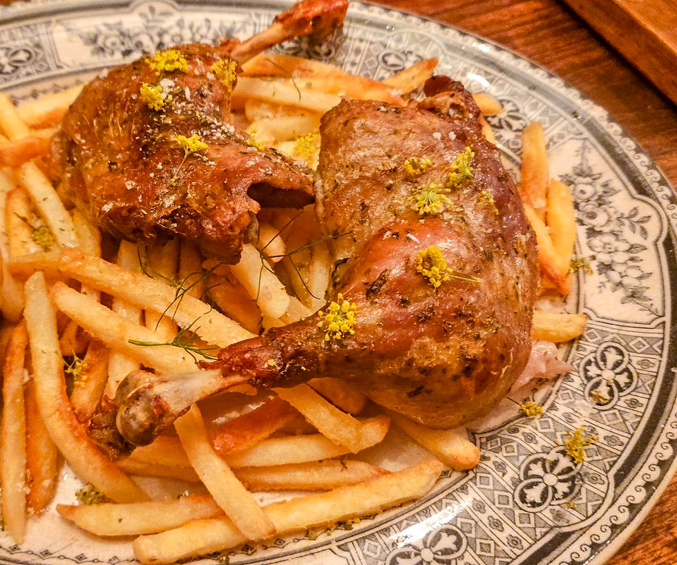 Roasted duck legs with fries on a patterned plate, garnished with yellow herbs. The scene is warm and appetizing.