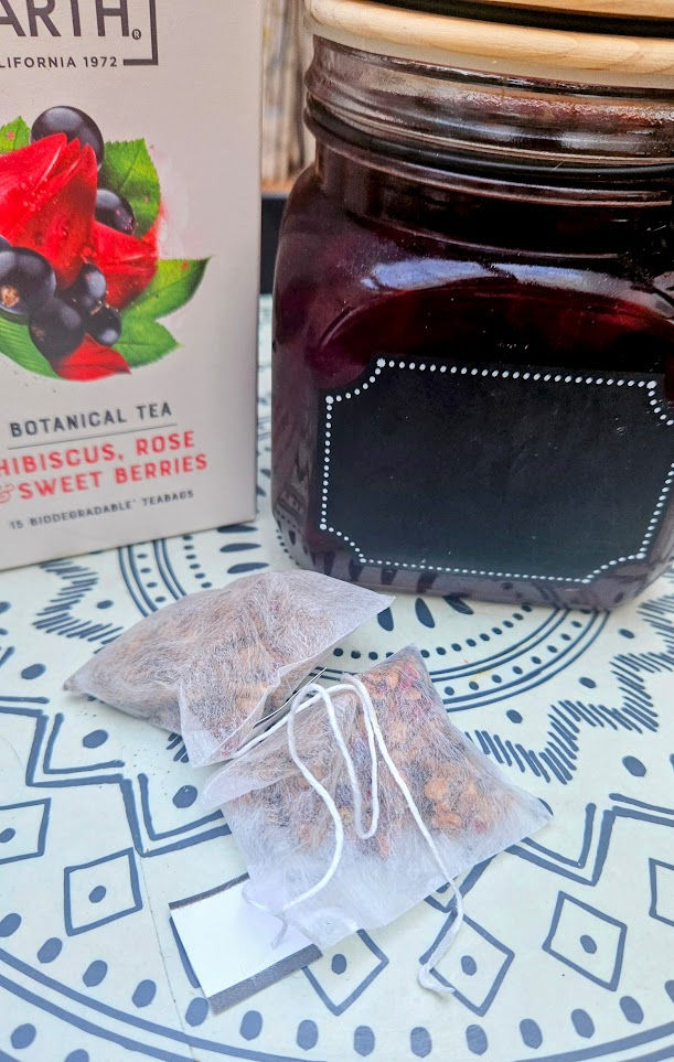 Tea bags on a patterned table next to a jar with a blank label and a box labeled Hibiscus, Rose & Sweet Berries Botanical Tea.