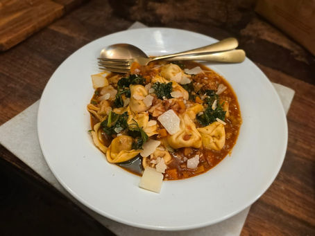 Plate of tortellini in brown sauce with greens and shaved cheese, topped with a spoon and fork on a wooden table.
