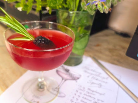 Pink cocktail in a coupe with blackberry and rosemary, resting on a floral menu beside a vase of herbs and flowers.