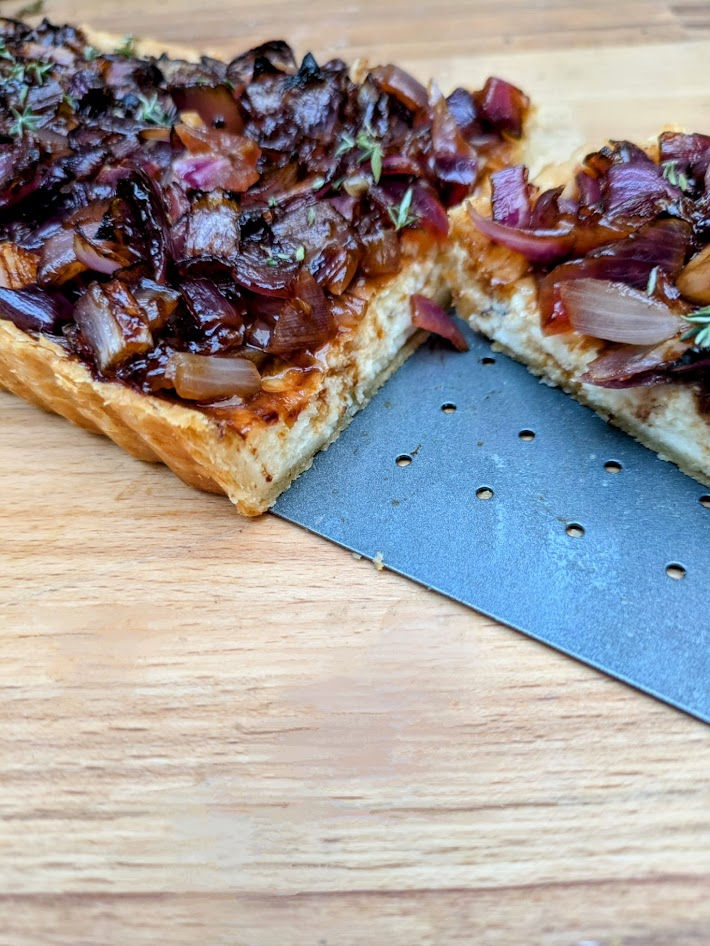 Caramelized onion tart on wooden board, topped with thyme. A slice is cut, revealing flaky crust. Perforated spatula visible.