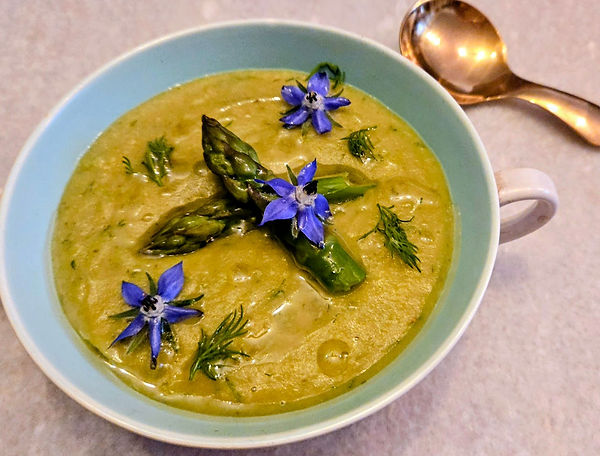 Green soup in a pale blue bowl garnished with asparagus tips and blue borage flowers