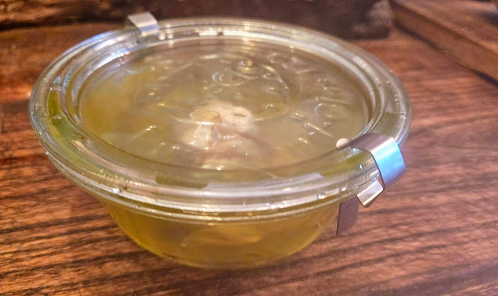 A glass jar with a metal clasp contains green pickles, set on a wooden surface. The lid shows faint impressions of text. Warm lighting.