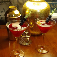 2 martini glasses filled with a red liquid, garnished with blackberries on a rosemary spring, sat on a dark wooden surface