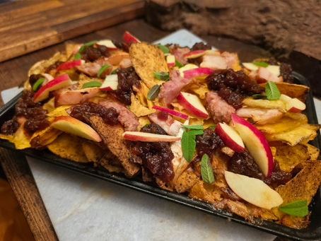 Loaded nachos topped with shredded meat, cheese, apple slices, red onion, and herbs on a black tray in a rustic setting.