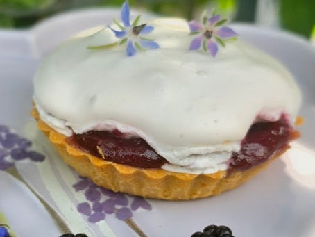 White-meringue topped berry tart with red filling and edible flower garnish on a floral plate, with two blackberries nearby.