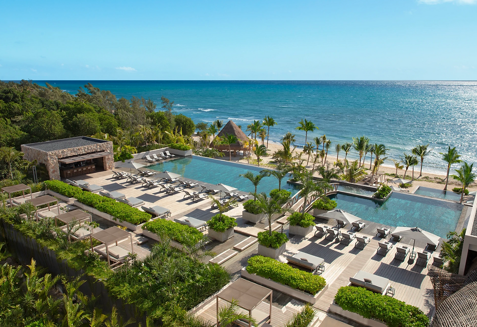 An elevated, high-angle view of a luxury oceanfront resort's expansive pool deck. Two pristine infinity pools are surrounded by light-colored wooden decking lined with modern lounge chairs and gray umbrellas. Lush green manicured hedges, tropical palm trees, and an open-air stone pool bar flank the deck. In the background, a white-sand beach with a thatched-roof palapa leads to a vibrant turquoise ocean under a clear blue sky.