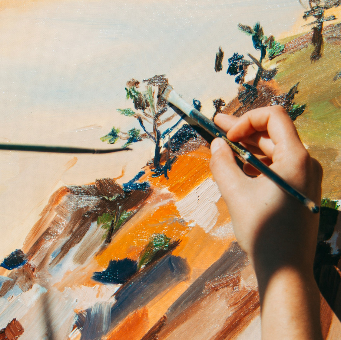painters hand painting a picture of a landscape with trees and hills in orange and green hues