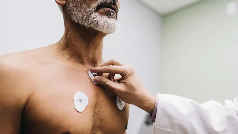 A person places electrodes on an older man's chest in a medical setting. The background is neutral, with a calm atmosphere.