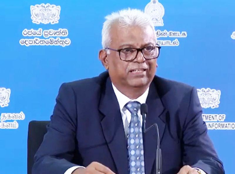 Man in glasses speaking at a podium, wearing a suit and tie. Blue backdrop with logos and text reads "Department of Information."