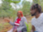 Two Black people one older one younger are sitting outside on a slope of red earth with green coffee bushes behind them.