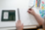 A close-up of the designer's hands drawing an intricate, hand-drawn pattern in a sketchbook using a green Faber-Castell pencil. A set of green Faber-Castell pencils is neatly arranged in an open tin beside the sketchbook. A watercolour palette with vibrant colours and a separate sheet of blue and green watercolour swatches are also visible on the desk.