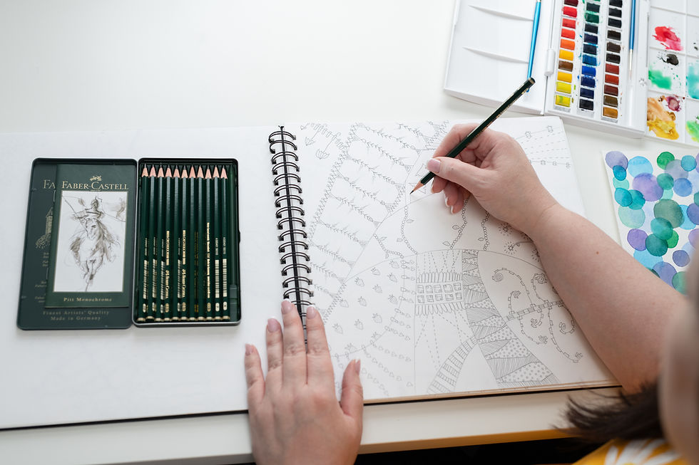 A close-up of the designer's hands drawing an intricate, hand-drawn pattern in a sketchbook using a green Faber-Castell pencil. A set of green Faber-Castell pencils is neatly arranged in an open tin beside the sketchbook. A watercolour palette with vibrant colours and a separate sheet of blue and green watercolour swatches are also visible on the desk.