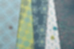The image shows a collection of fabric samples arranged in overlapping layers. Each fabric features Jo Phillip's pattern designs of intricate, repeating geometric and floral patterns in a harmonious colour palette of soft blues, greens, and teals. Some of the fabrics are accented with buttons in matching colours, adding texture and a touch of dimension to the display. The designs evoke a sense of delicate symmetry and organic flow, reminiscent of mandalas or lacework.