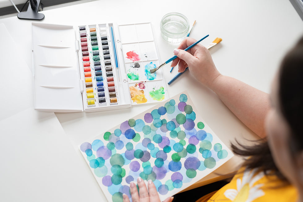 Jo Phillips, surface pattern designer, painting colourful overlapping circles in shades of blue, green, and purple using watercolours. A vibrant paint palette, mixing tray, and brushes are arranged on a white desk, with a glass of water nearby. Jo is wearing a yellow top with a white daisy print.