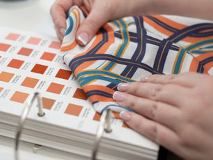 Close-up of hands holding a printed fabric swatch against a Pantone colour guide, comparing orange tones for pattern and textile matching