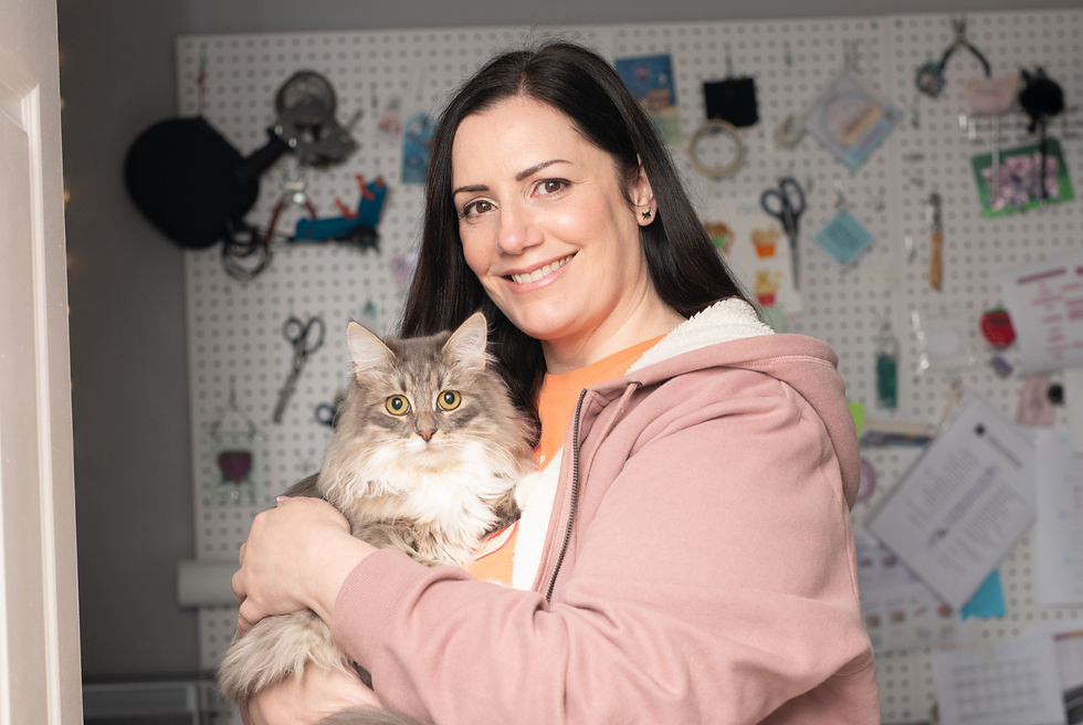 Jo has dark hair and wears a pink hoodie. She stands in her studio smiling and holding her fluffy cat, Monty