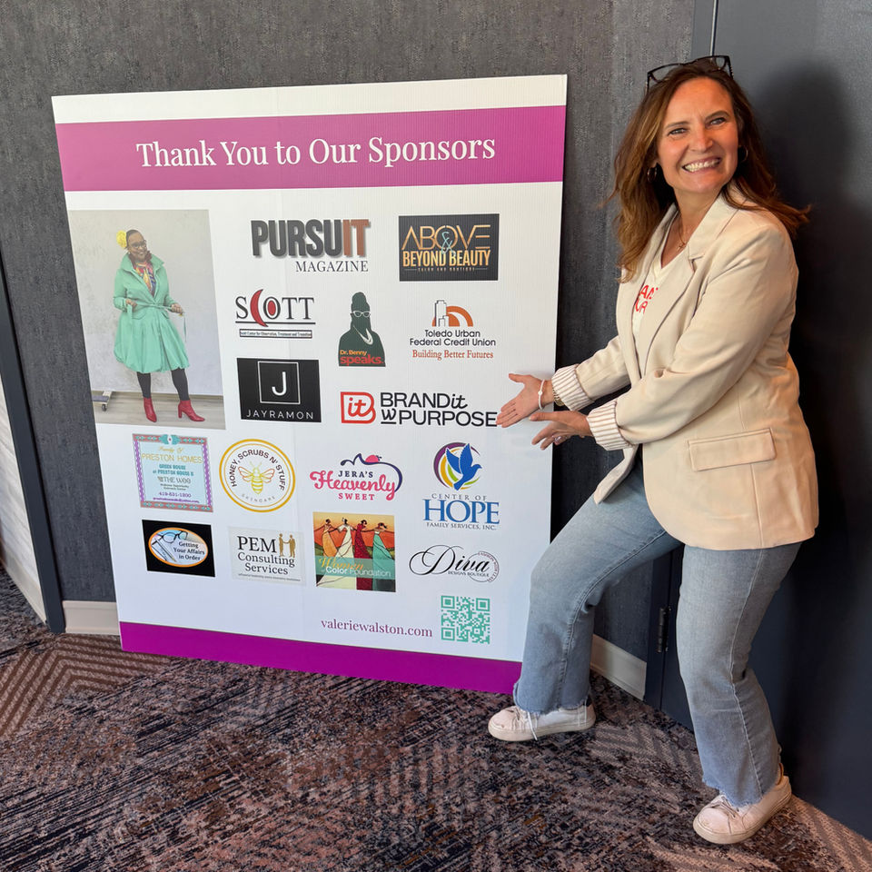 Woman smiling and pointing to a sponsor recognition board featuring local business and nonprofit logos at a community event.