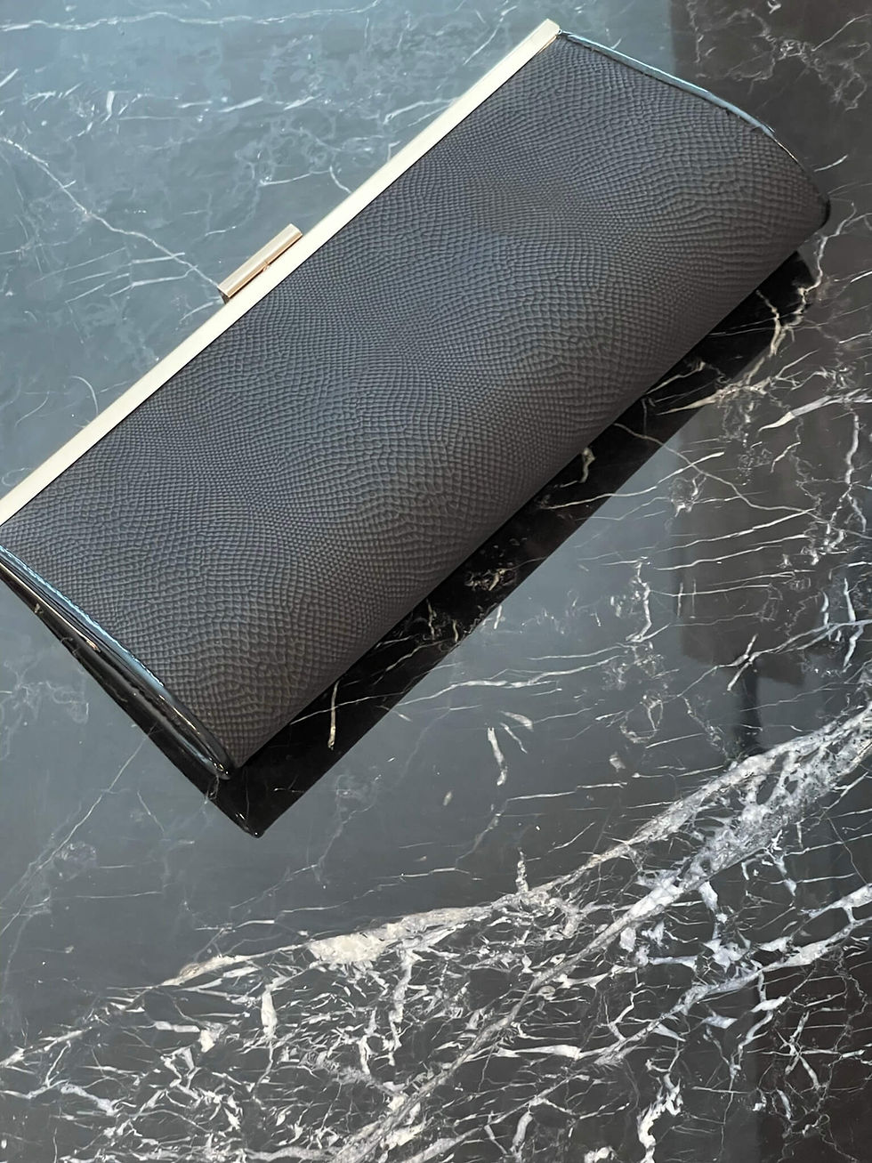 New Look Textured Clutch – angled top view showing black textured surface and gold-tone frame on black marble top