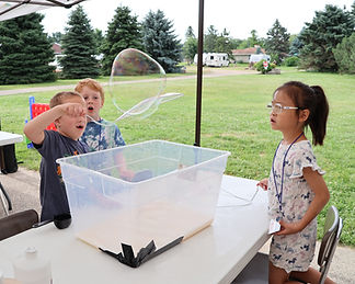 Three smiling students squat close to the ground and swing cups containing their handmade liquid chalk on a pendulum to create a design on paper laying on the ground.