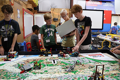 Three students stand at the robot game table with their laptop and LEGO robot preparing to run a robot mission.