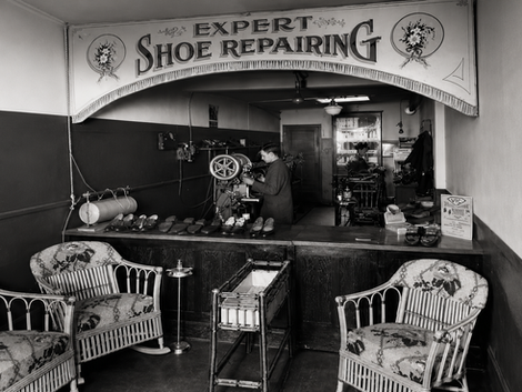 Old shoe repair shop: 'Expert Shoe Repairing' sign, cobbler at work.