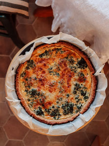Spinach and cheese quiche in a baking dish on a wooden surface, with a light cloth and patterned chair in the background. Warm, inviting setting.