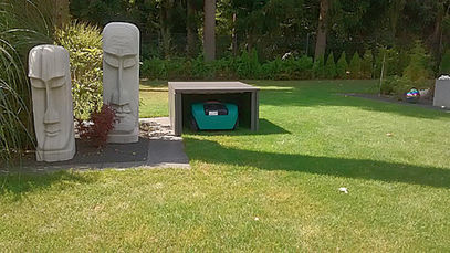 Zwei abstrakte Steinskulpturen und Rasenroboter-Garage in einem grünen Garten.